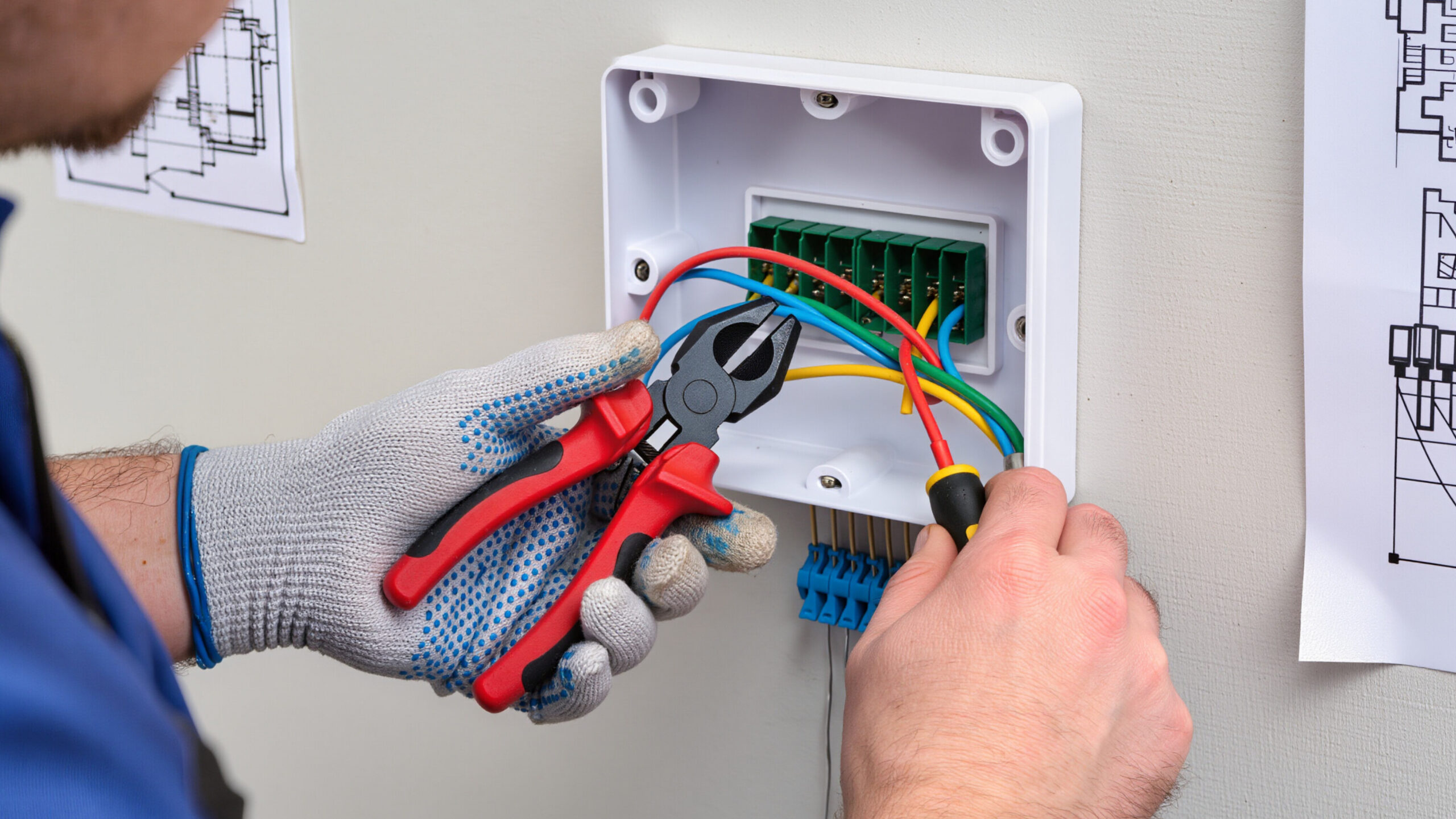 Electrician connecting wires in electrical panel during whole-home rewire, with tools and blueprints visible.